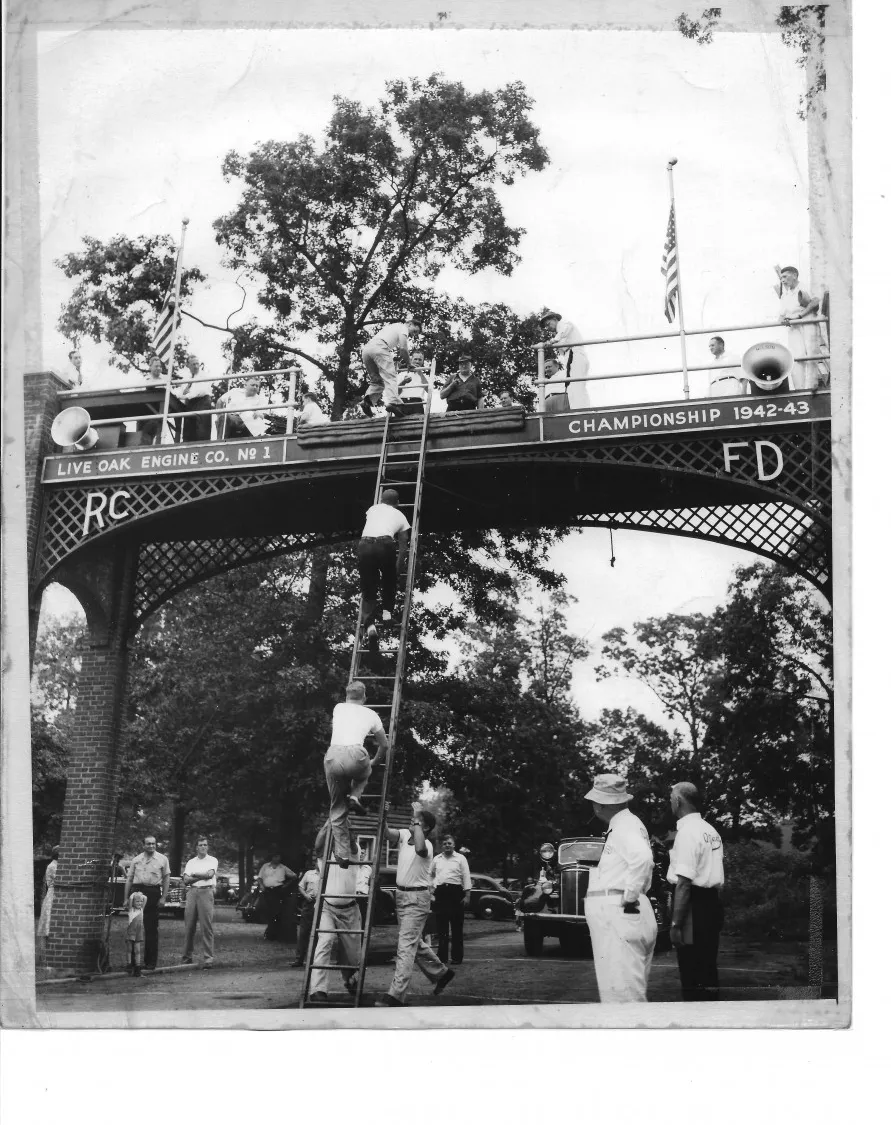 Historic photograph of firefighters using ladders at the Rockville Centre firemen's tournament arch.